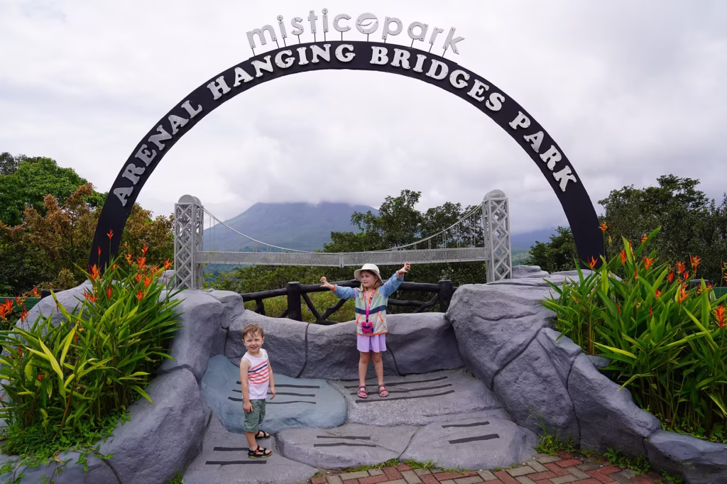 2 young kids standing and smiling in front of the misticopark entrance sign with Arenal volcano in the background in Costa Rica.
